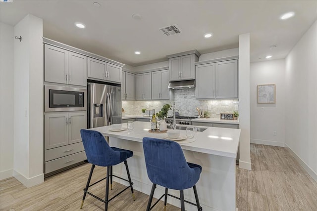 kitchen featuring built in microwave, stainless steel fridge with ice dispenser, gray cabinets, a kitchen island with sink, and a kitchen bar