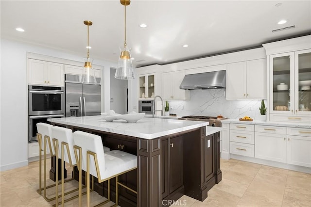 kitchen with a center island with sink, appliances with stainless steel finishes, hanging light fixtures, wall chimney exhaust hood, and light stone counters