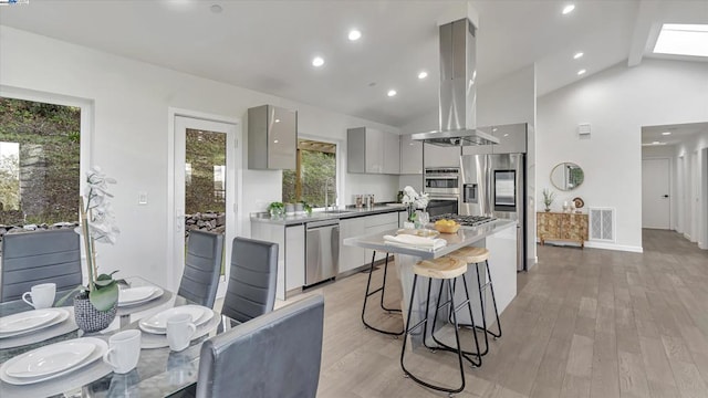 kitchen with gray cabinetry, island exhaust hood, stainless steel appliances, lofted ceiling with beams, and light hardwood / wood-style flooring