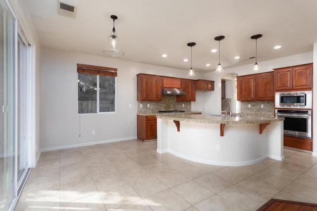 kitchen with appliances with stainless steel finishes, a breakfast bar, tasteful backsplash, light stone counters, and a center island with sink