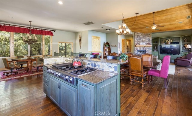 kitchen featuring stainless steel gas stovetop, a stone fireplace, an island with sink, decorative light fixtures, and light stone counters