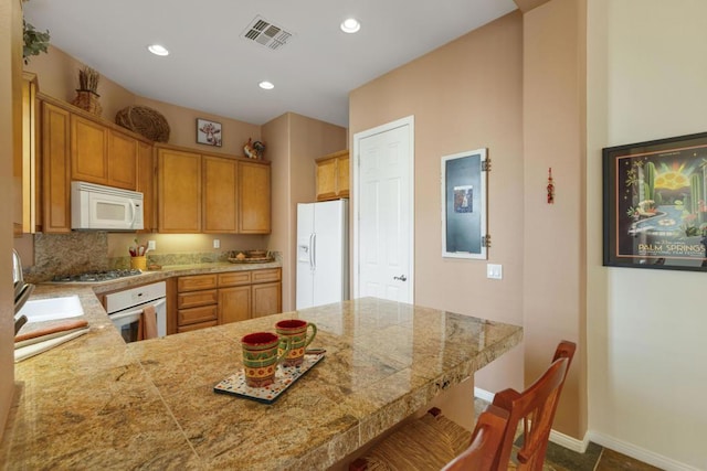 kitchen featuring backsplash, dark tile patterned flooring, kitchen peninsula, white appliances, and a kitchen breakfast bar