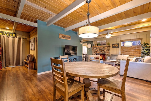 dining room with hardwood / wood-style floors, beamed ceiling, a stone fireplace, wooden walls, and ceiling fan