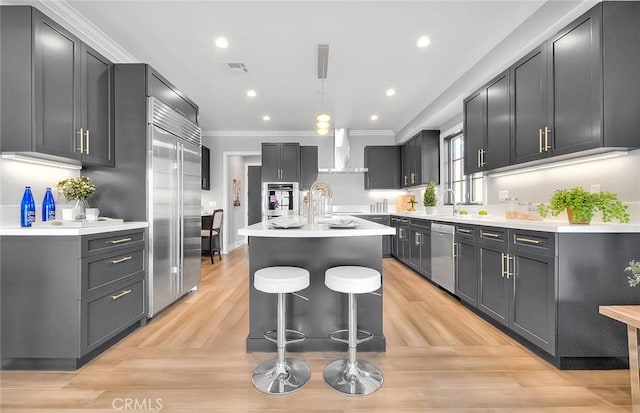 kitchen with an island with sink, appliances with stainless steel finishes, decorative light fixtures, wall chimney range hood, and crown molding