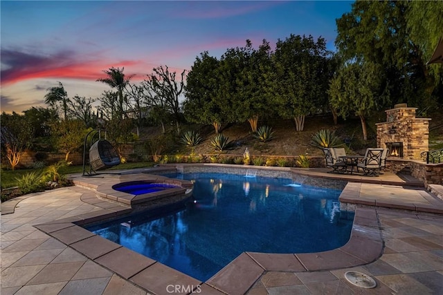 pool at dusk featuring an in ground hot tub, a patio, and an outdoor stone fireplace