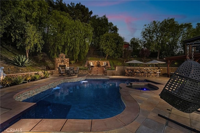 pool at dusk featuring an outdoor stone fireplace, an in ground hot tub, and a patio