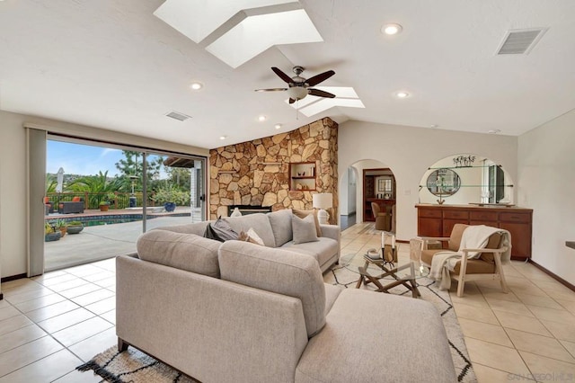 living room with ceiling fan, light tile patterned floors, a stone fireplace, and vaulted ceiling with skylight