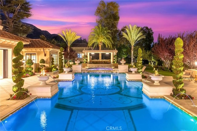 pool at dusk featuring an in ground hot tub, a patio area, an outdoor structure, and pool water feature