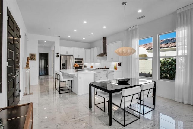 kitchen featuring an island with sink, a breakfast bar area, white cabinets, stainless steel appliances, and wall chimney range hood