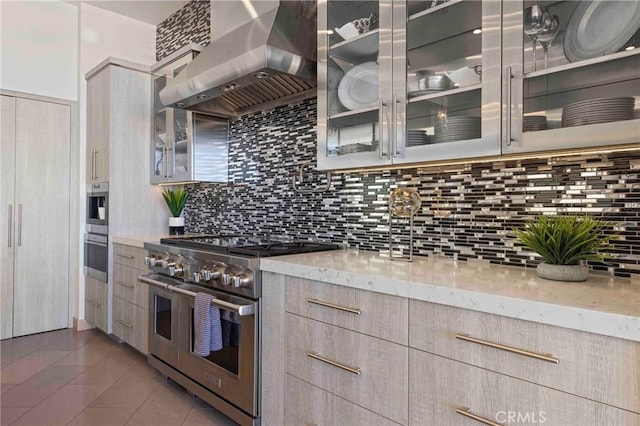 kitchen featuring decorative backsplash, light brown cabinets, range with two ovens, wall chimney exhaust hood, and light stone counters