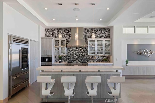 kitchen with a large island with sink, pendant lighting, wall chimney exhaust hood, stainless steel appliances, and a kitchen breakfast bar