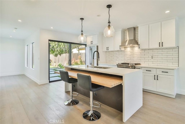 kitchen featuring a kitchen island with sink, hanging light fixtures, wall chimney exhaust hood, white cabinets, and sink