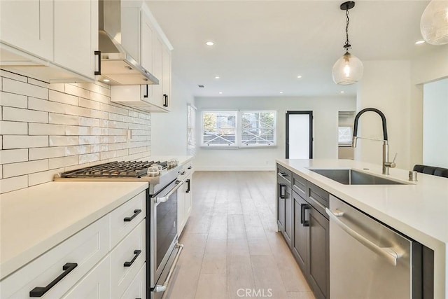 kitchen featuring hanging light fixtures, stainless steel appliances, wall chimney range hood, white cabinetry, and sink