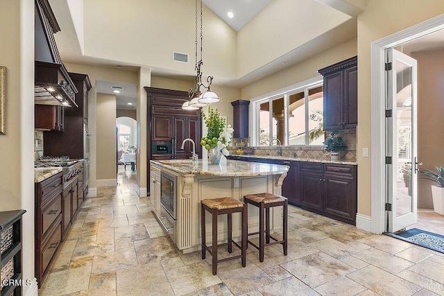 kitchen with stainless steel gas stovetop, custom exhaust hood, an island with sink, pendant lighting, and light stone counters