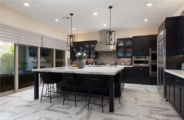 kitchen featuring an island with sink, a breakfast bar area, decorative backsplash, decorative light fixtures, and wall chimney exhaust hood