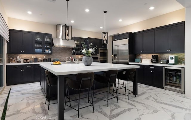 kitchen featuring beverage cooler, appliances with stainless steel finishes, wall chimney exhaust hood, hanging light fixtures, and a kitchen island with sink