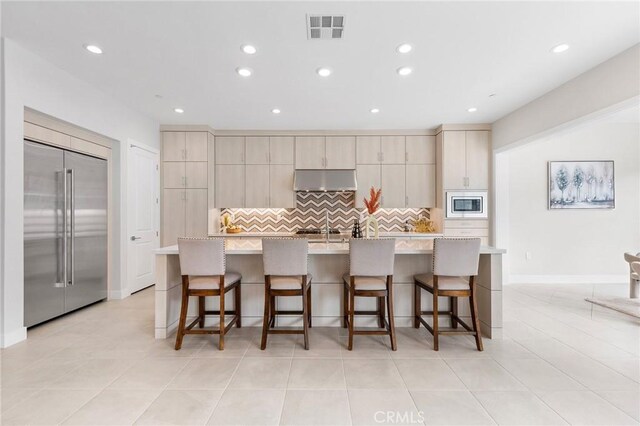 kitchen featuring ventilation hood, a kitchen island with sink, built in appliances, and a breakfast bar area