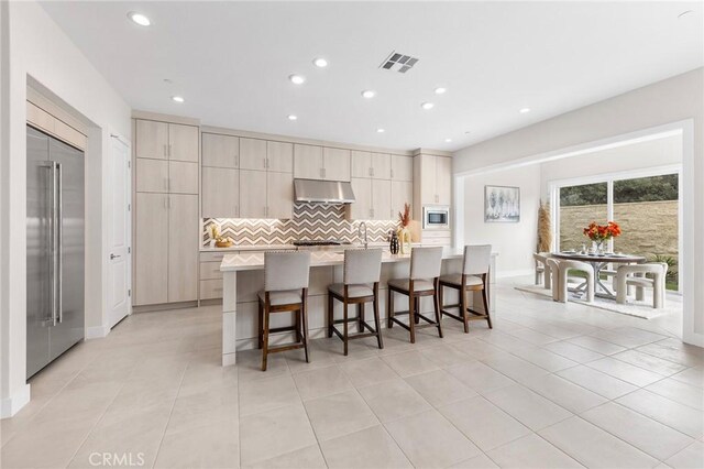 kitchen featuring backsplash, built in appliances, an island with sink, a breakfast bar area, and light tile patterned floors