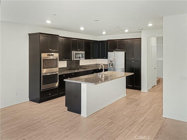 kitchen featuring light stone counters, light wood-type flooring, stainless steel appliances, and a center island with sink