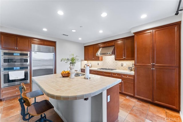 kitchen featuring extractor fan, tile countertops, a kitchen bar, a kitchen island with sink, and stainless steel appliances