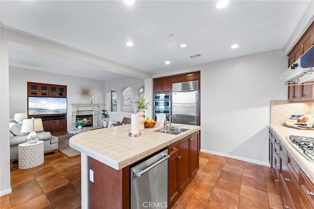 kitchen with extractor fan, an island with sink, stainless steel appliances, tile counters, and sink