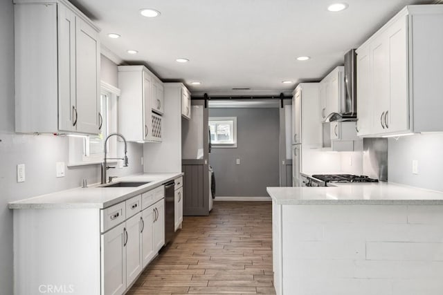 kitchen with a barn door, dishwasher, sink, white cabinetry, and wall chimney exhaust hood