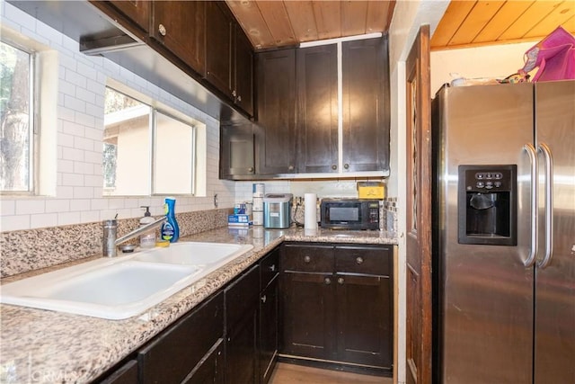 kitchen featuring light stone countertops, backsplash, dark brown cabinetry, sink, and stainless steel fridge with ice dispenser