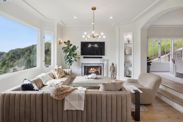 living room with built in shelves, a fireplace, a notable chandelier, light wood-type flooring, and crown molding