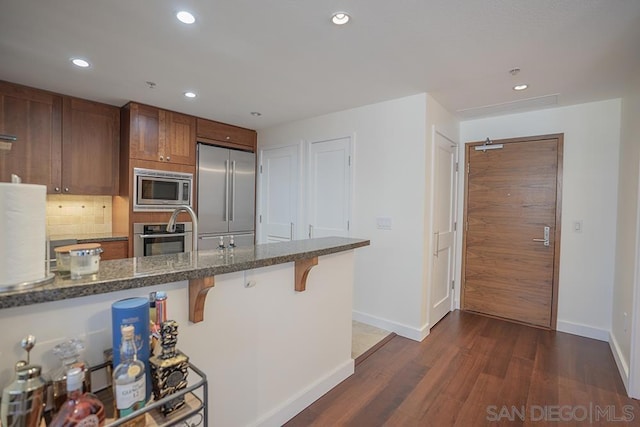 kitchen with appliances with stainless steel finishes, dark wood-type flooring, dark stone counters, decorative backsplash, and a breakfast bar