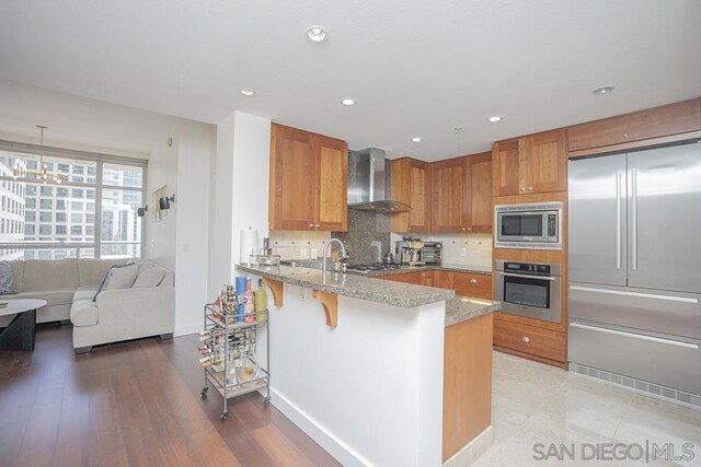 kitchen with decorative light fixtures, wall chimney range hood, built in appliances, kitchen peninsula, and a breakfast bar area