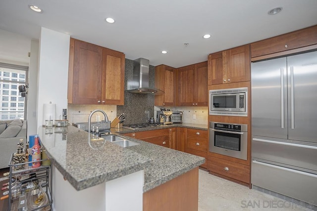 kitchen featuring wall chimney range hood, built in appliances, sink, stone countertops, and kitchen peninsula