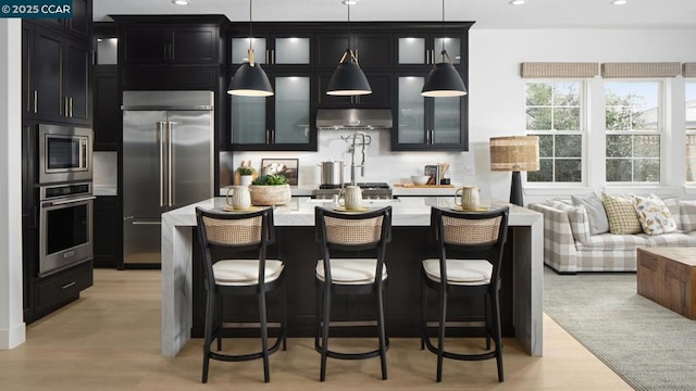 kitchen with an island with sink, light stone counters, hanging light fixtures, and built in appliances