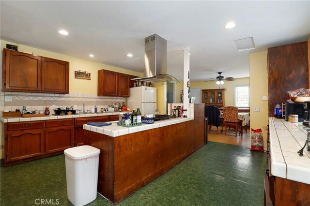 kitchen featuring tile countertops, ceiling fan, white fridge, kitchen peninsula, and island exhaust hood