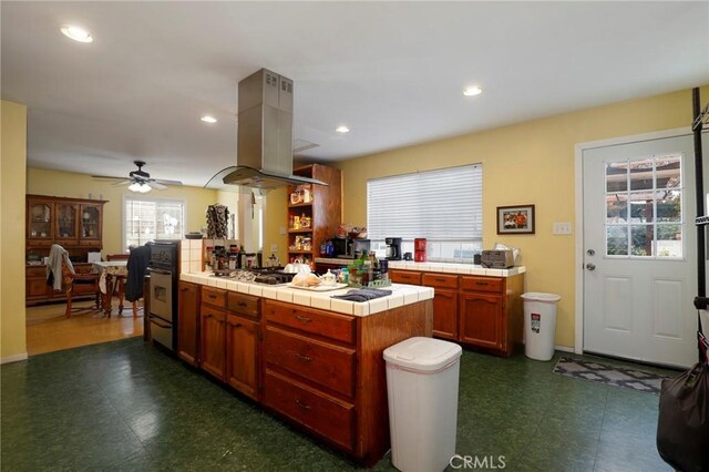 kitchen featuring tile countertops, a center island, ceiling fan, island range hood, and stainless steel gas cooktop