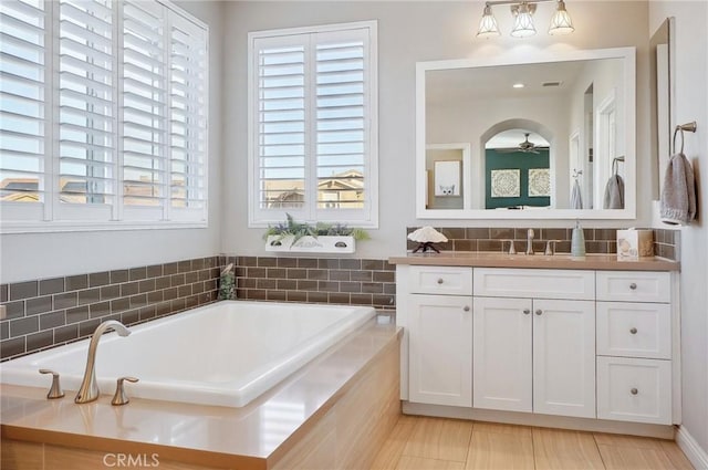 bathroom featuring ceiling fan, a relaxing tiled tub, and vanity