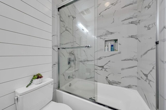 bathroom featuring toilet, combined bath / shower with glass door, and wood walls