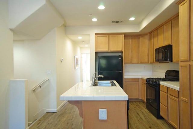 kitchen featuring black appliances, light hardwood / wood-style floors, tile counters, a kitchen island with sink, and sink