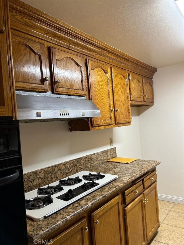 kitchen featuring under cabinet range hood, light tile patterned flooring, dark countertops, white gas stovetop, and baseboards