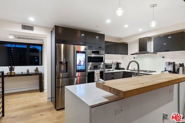 kitchen with appliances with stainless steel finishes, wood counters, decorative light fixtures, wall chimney range hood, and light hardwood / wood-style flooring