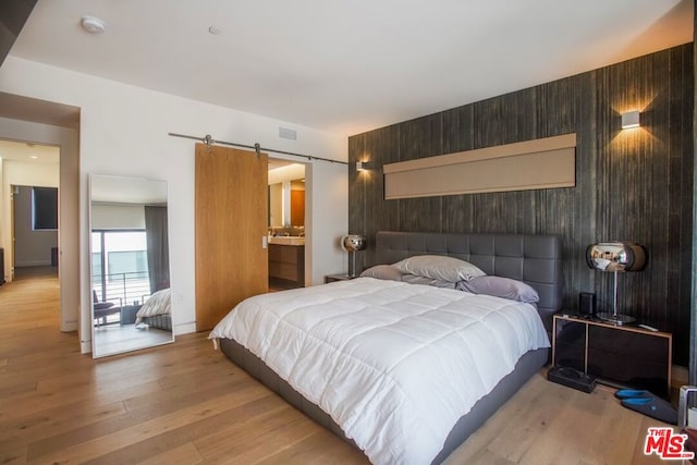 bedroom featuring ensuite bath, a barn door, wooden walls, and light hardwood / wood-style flooring