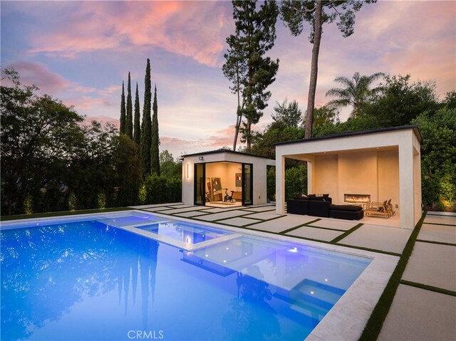 pool at dusk featuring an in ground hot tub, an outdoor structure, an outdoor hangout area, and a patio