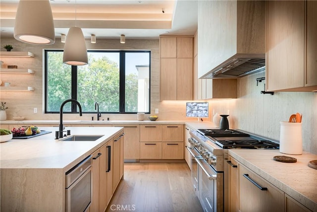 kitchen featuring double oven range, light brown cabinetry, hanging light fixtures, wall chimney range hood, and sink