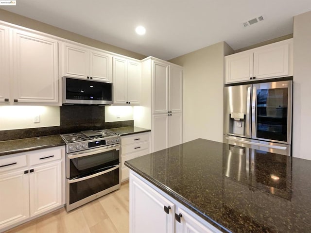 kitchen with appliances with stainless steel finishes, white cabinetry, dark stone counters, tasteful backsplash, and light hardwood / wood-style flooring