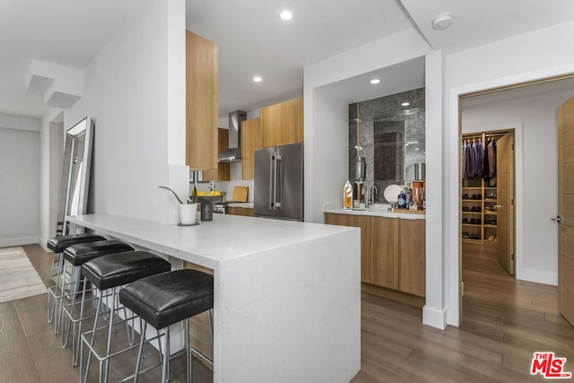 kitchen with high end fridge, a breakfast bar area, dark wood-type flooring, wall chimney exhaust hood, and sink