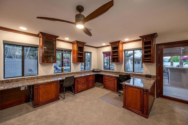 kitchen with ceiling fan, light colored carpet, dark stone countertops, built in desk, and crown molding