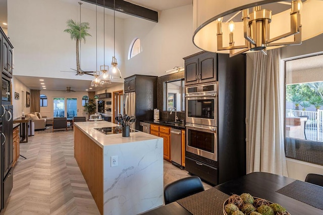 kitchen with ceiling fan with notable chandelier, stainless steel appliances, beamed ceiling, an island with sink, and sink