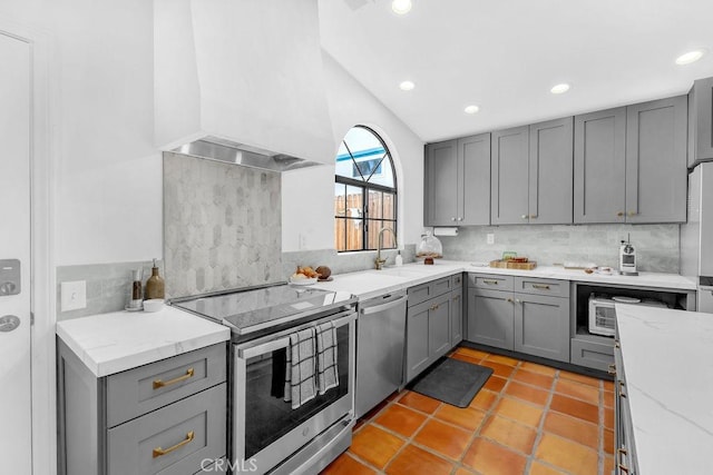 kitchen with lofted ceiling, custom exhaust hood, gray cabinetry, stainless steel appliances, and light stone counters