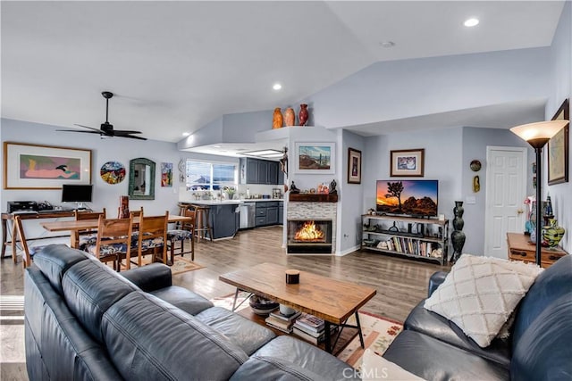 living room featuring ceiling fan, light hardwood / wood-style flooring, a stone fireplace, and vaulted ceiling