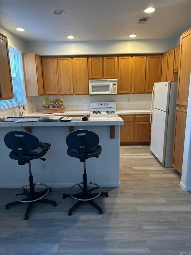 kitchen featuring kitchen peninsula, tile counters, white appliances, a kitchen breakfast bar, and light hardwood / wood-style flooring