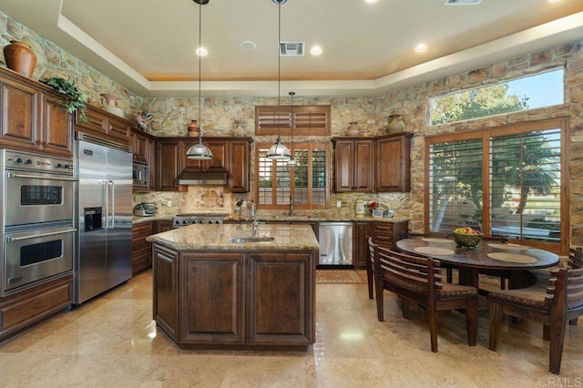 kitchen with light stone counters, built in appliances, a sink, under cabinet range hood, and backsplash